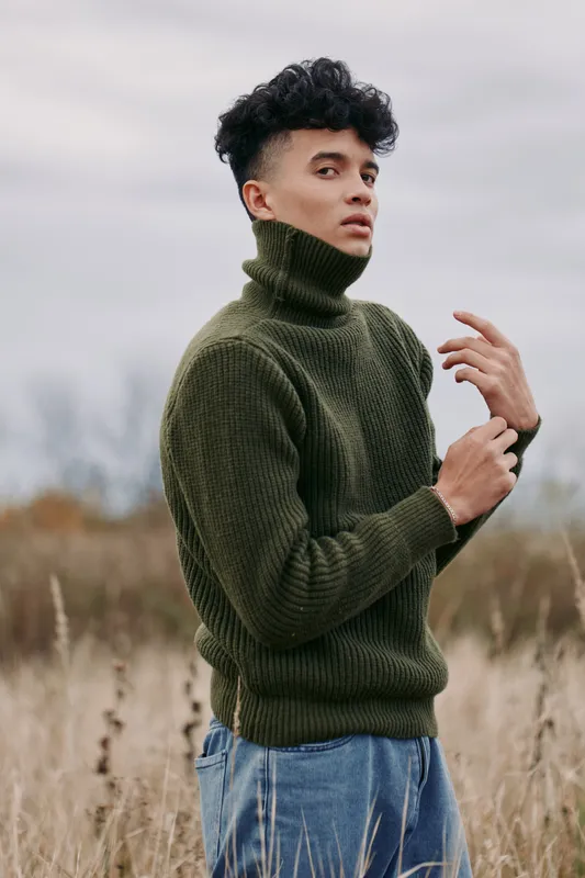Portrait of a young man wearing a green turtleneck sweater in a natural field, captured with authentic, credible emotion and subtle gaze, emphasizing genuine mood and texture.