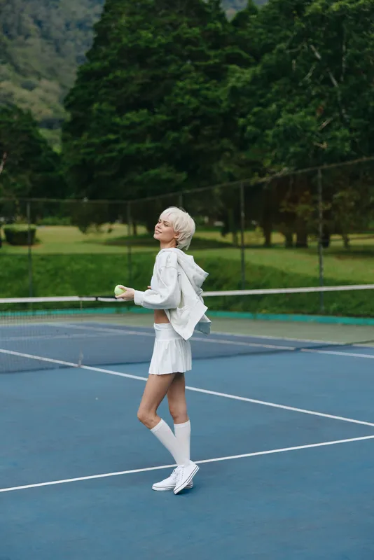 Young woman in a stylish tennis outfit, smiling on a tennis court, surrounded by greenery, representing a sporty, vibrant, and healthy lifestyle