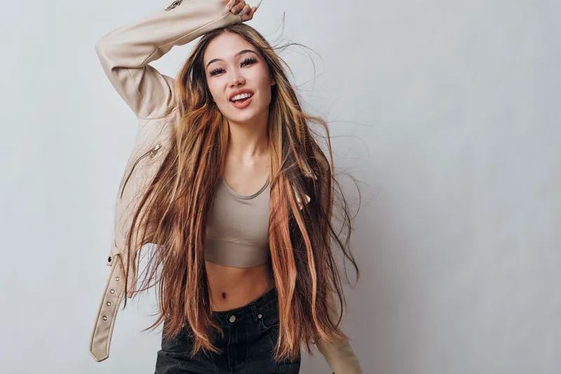 Young woman with long hair posing joyfully and smiling in casual beige top and jacket against light background for portrait and lifestyle concept.