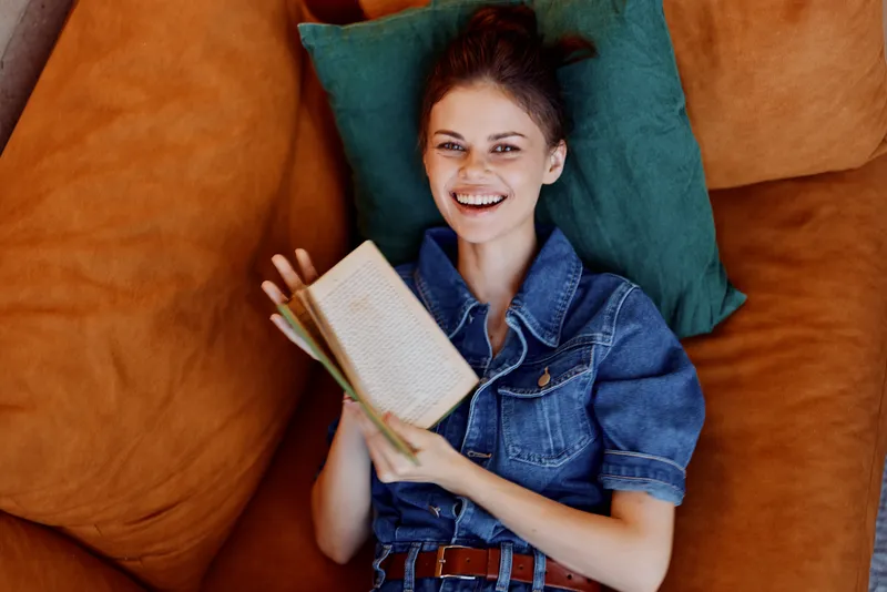 Счастливая женщина отдыхает на диване с книгой в руке и мирно улыбается