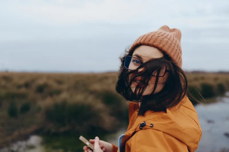 Woman lifestyle outdoors winter hat scarf mask smartphone river landscape, wearing orange coat, holding smartphone, looking at camera, calm mood, natural daylight
