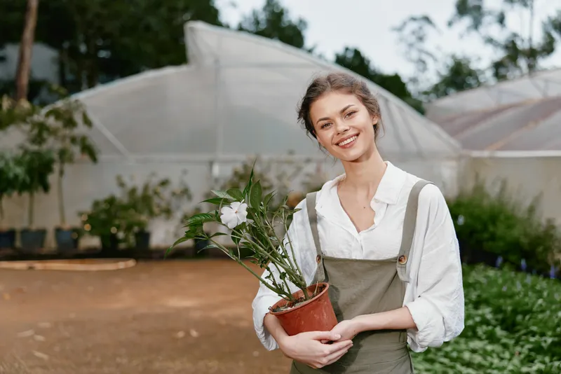 Young woman in an apron smiling while holding a potted flower, showcasing a sustainable gardening concept in a greenhouse environment