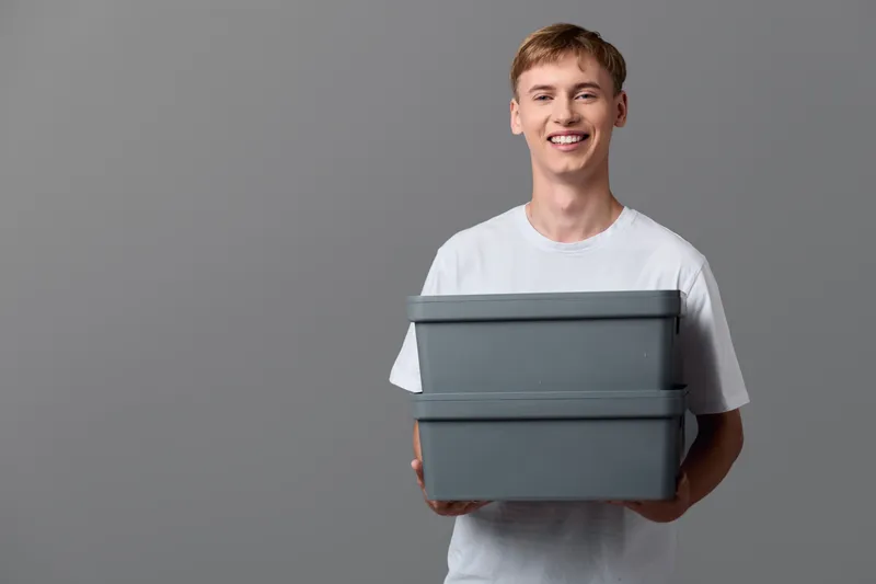Man holding gray boxes in a white T shirt against a neutral studio backdrop, illustrating packaging, storage and delivery for ecommerce order processing and logistics efficiency