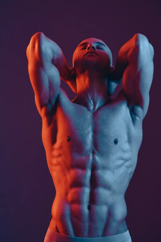 Muscular man poses dramatically with hands behind his head, showcasing a strong physique in a stunning light composition. High contrast colors create an engaging atmosphere.