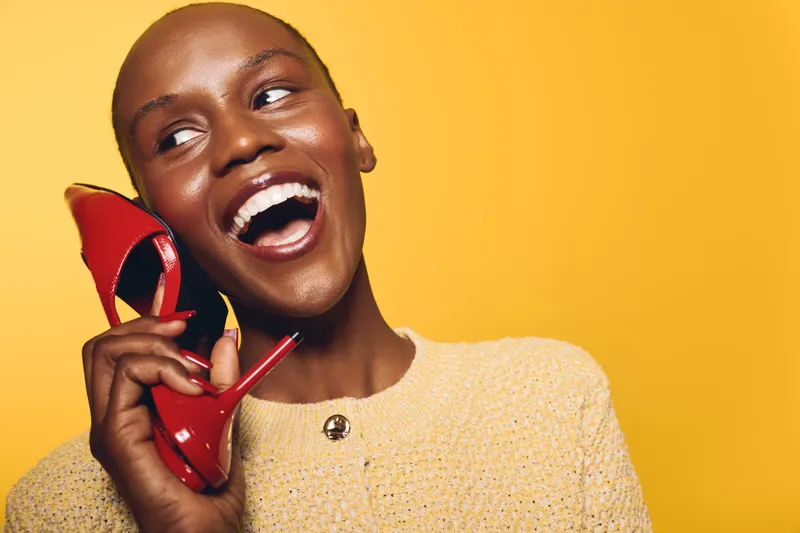Happy black woman with dark skin holds red high heel shoe near face, laughing and looking away on yellow background. Concept for fashion, joy, and beauty with vibrant colors.