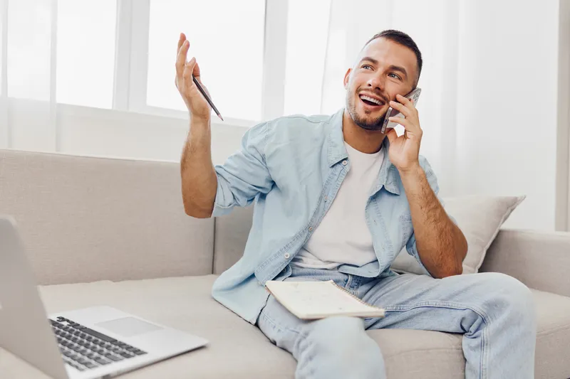 Happy man talking on his phone while sitting on a sofa, dressed casually, expressing joy and enthusiasm in a bright, airy setting with natural light.