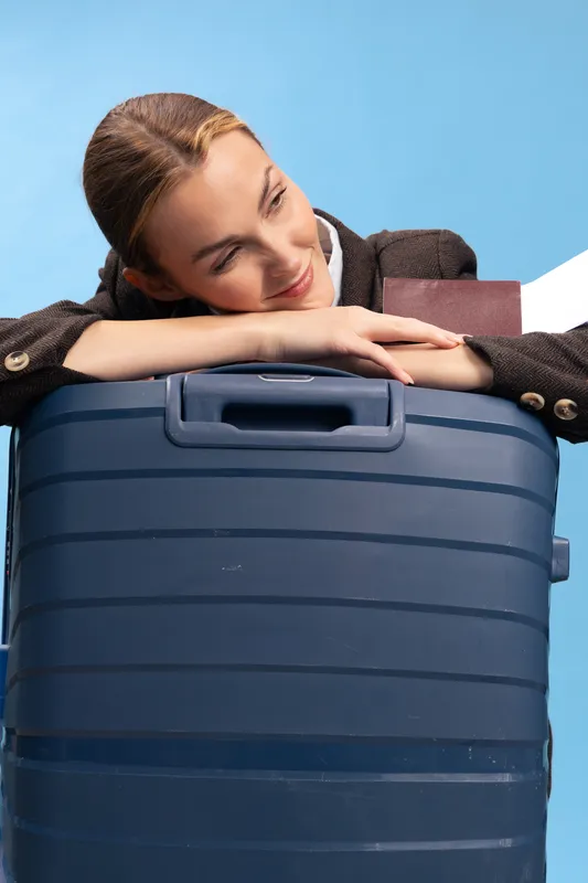 Young woman in brown jacket resting her head on large blue suitcase, smiling with closed eyes on light blue background. Concept of travel, vacation, and relaxation before trip.