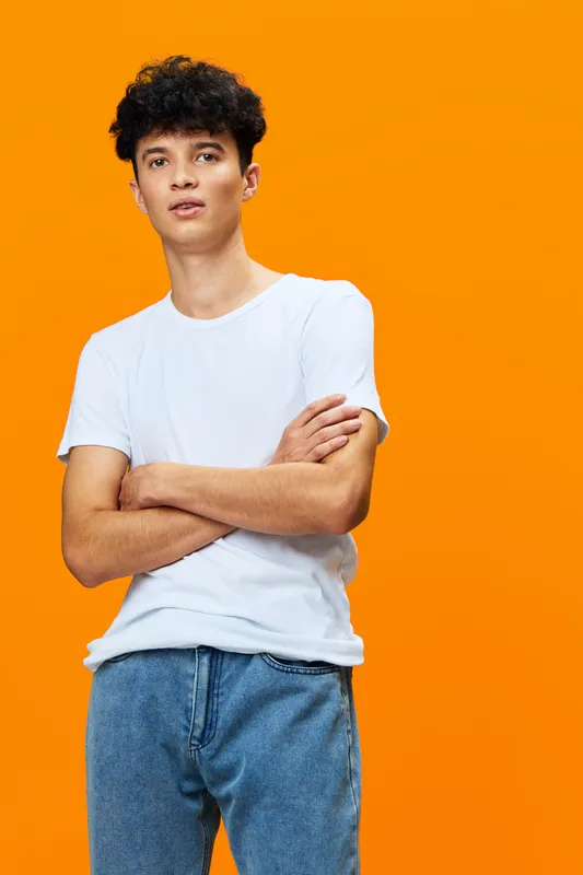 young man with curly hair wearing white t-shirt and blue jeans stands with crossed arms against vibrant orange background indoors casual style expression