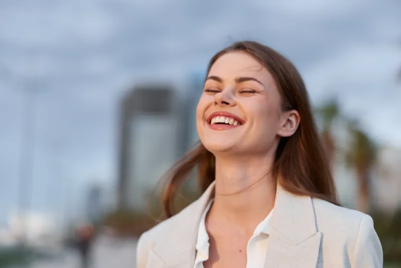 Happy woman smiling outdoors in casual light blazer during daytime with blurred city background, natural emotions and warm urban atmosphere