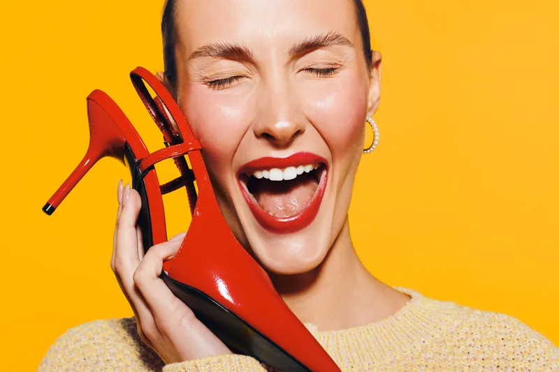 Happy woman with red lipstick holds red high heel shoe near face, eyes closed and mouth open. Bright yellow background and cozy sweater create vibrant, joyful fashion concept.