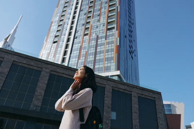Skyscraper skyline scene with a woman lifting her face toward the sun, backpack on her shoulder, exploring an urban landscape and admiring modern architecture, feeling hopeful
