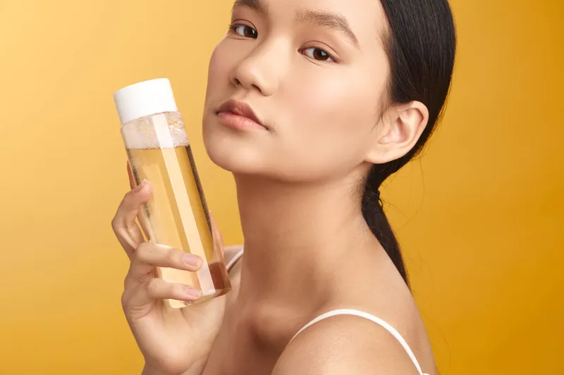 Asian woman holds a skincare product bottle against a warm backdrop, exuding calm confidence and a focused beauty routine in a modern studio setting.