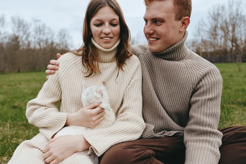 Happy couple outdoors with a goat kid in a cozy sweater moment, embracing family warmth in a field, a lifestyle candid scene under natural light.