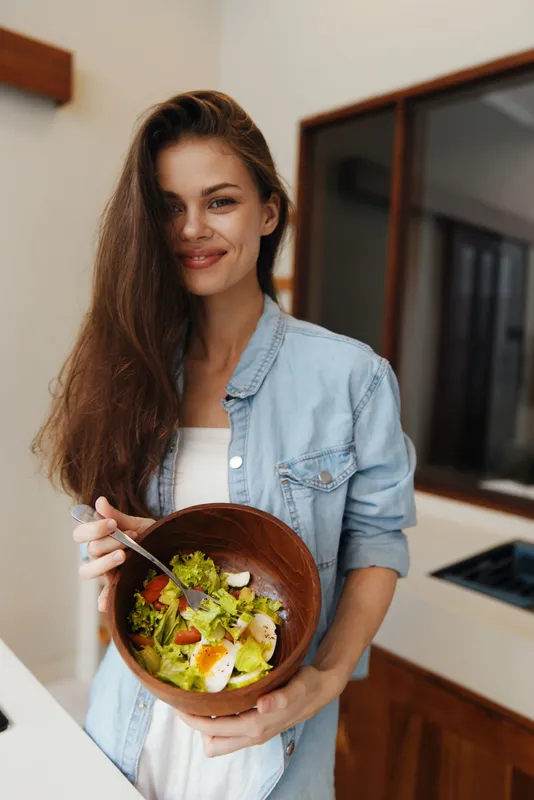 Весёлая молодая женщина в джинсовой куртке стоит на современной кухне, радостно держа деревянную миску с красочным салатом