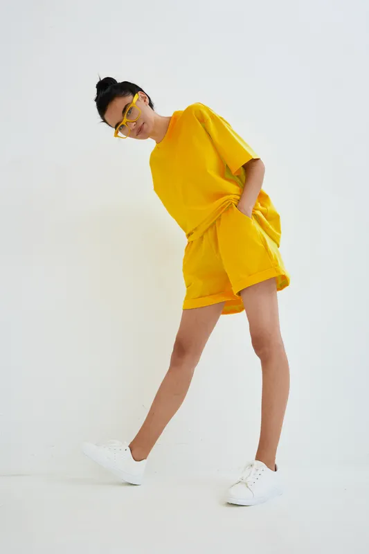 young woman in yellow summer outfit wearing matching sunglasses and white sneakers posing confidently against white background in casual style