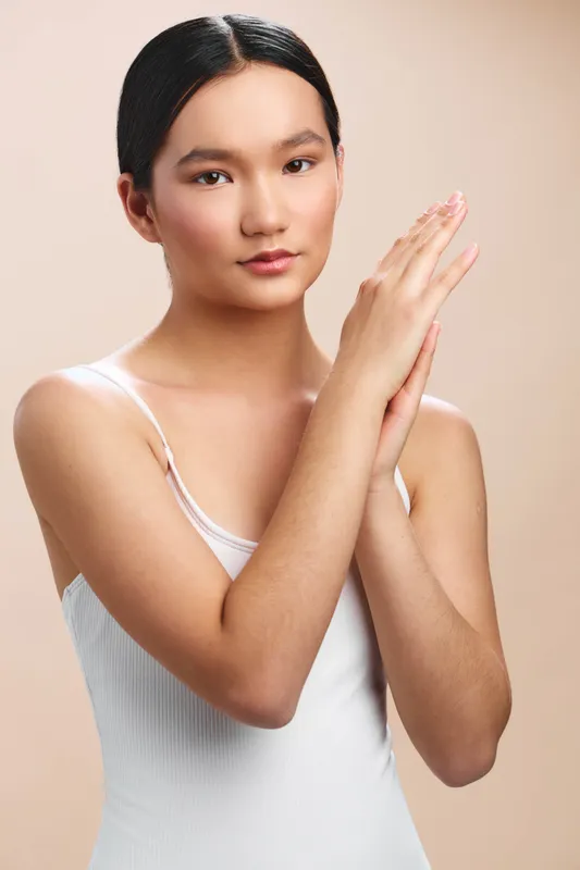 Asian woman in a white tank top stands against a warm neutral backdrop, her hands gently pressed together in a calm pose, conveying confidence, focus, and a serene studio mood.