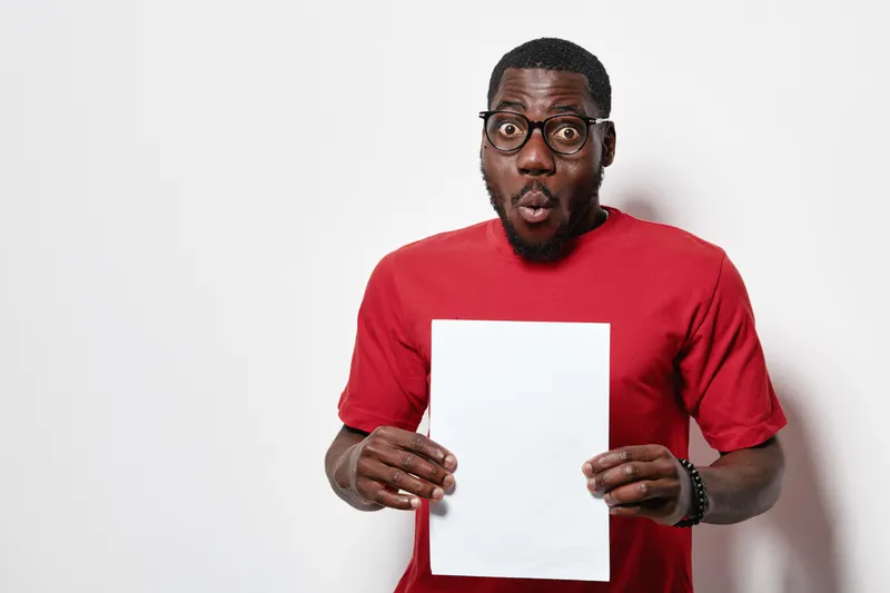 African American man with dark skin and glasses wearing red shirt holding blank white paper with surprised expression on white background.