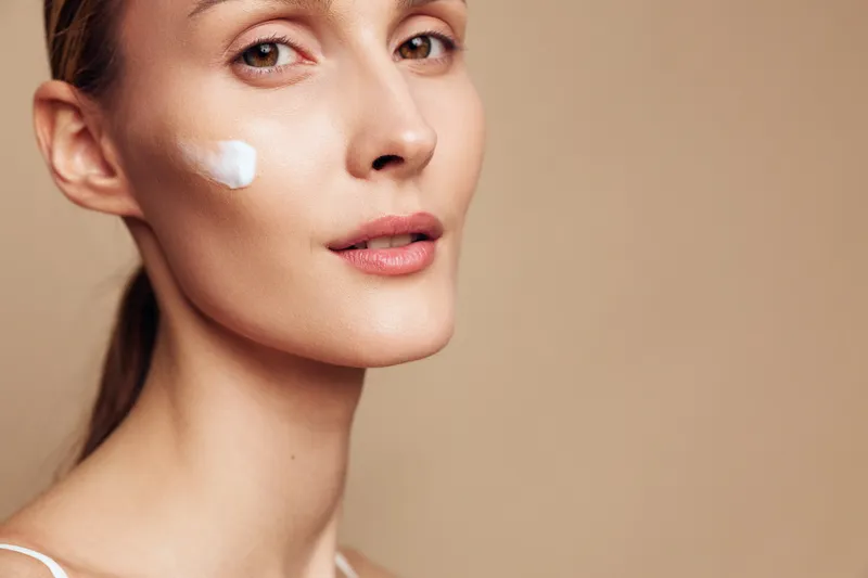 Woman applying face cream on cheek in a calm beauty close up, spa lighting, skincare and hydration concept, cosmetic treatment for healthy smooth skin, studio beige background