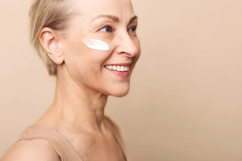 Smiling mature woman with short hair applies white cream on cheek, showing natural skin care routine and beauty. Soft beige background highlights healthy glow and self-care concept.