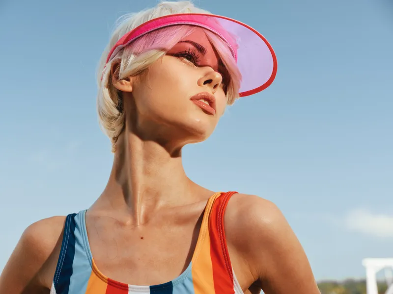 Portrait of a confident woman wearing a colorful striped swimsuit and pink visor under a clear blue sky, radiating summer energy and bold style.