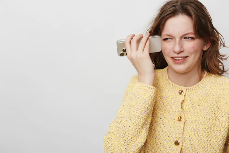 Young woman in yellow sweater holds smartphone to ear with confused expression on white background. Concept of misunderstanding, communication, and technology use.