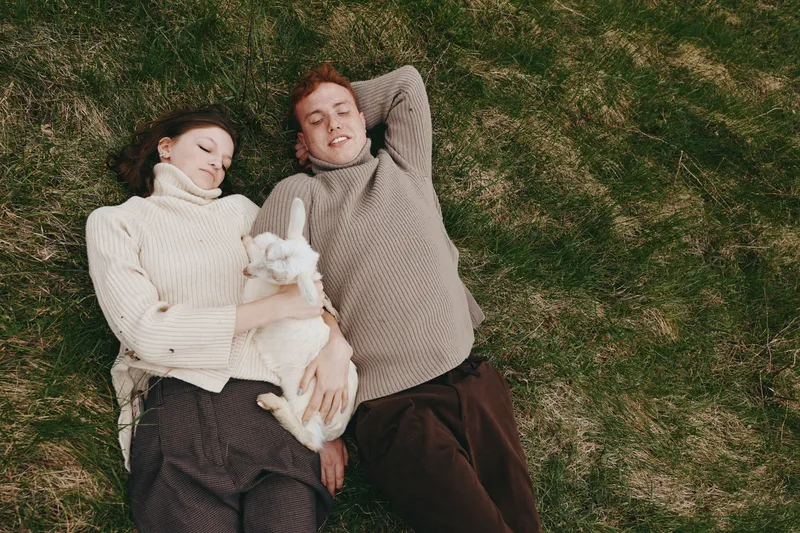 A couple outdoors on grass share a tender moment with a white goat kid, showcasing relaxed rural life, companionship and affection in a natural, intimate lifestyle scene, warmth.
