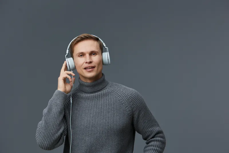 Young caucasian man wearing gray sweater and headphones listening and interacting with voice assistant for smart home technology on dark gray background studio portrait