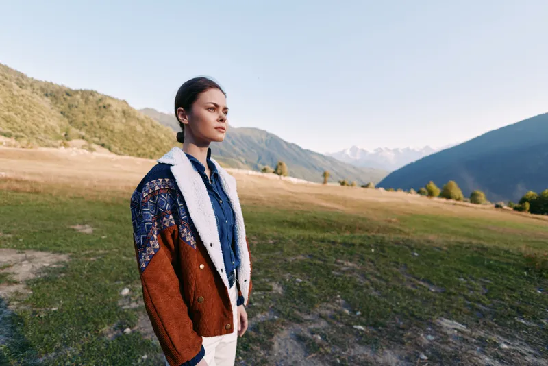 Woman portrait in mountains meadow outdoors nature, young model wearing knitted sweater and jacket standing in vast valley with thoughtful gaze and serene scenery at sunrise for travel lifestyle.