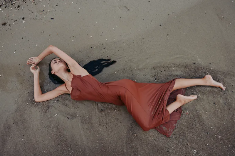 A solitary woman lies on sandy ground wearing a flowing red dress, creating a serene, artistic composition with soft shadows and a dramatic, graceful pose.
