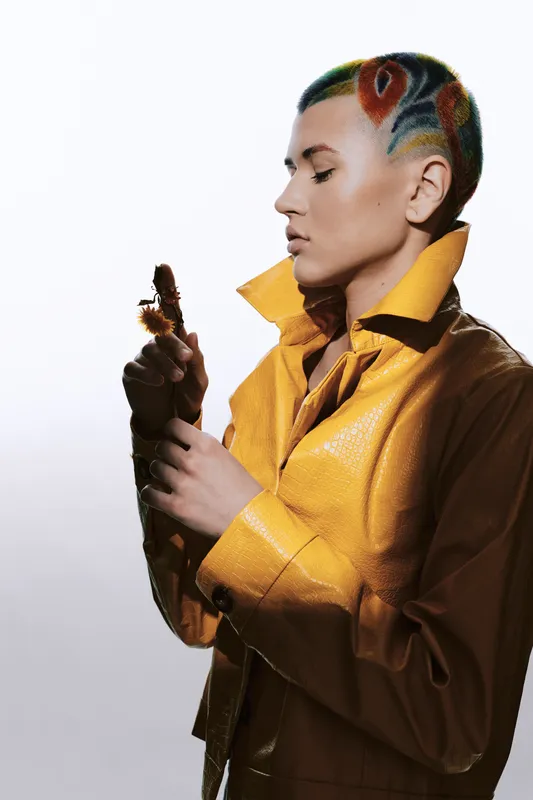 Woman with colorful patterned hair and yellow leather jacket holding a dried flower, showcasing modern style and thoughtful expression in a minimalistic studio setting.