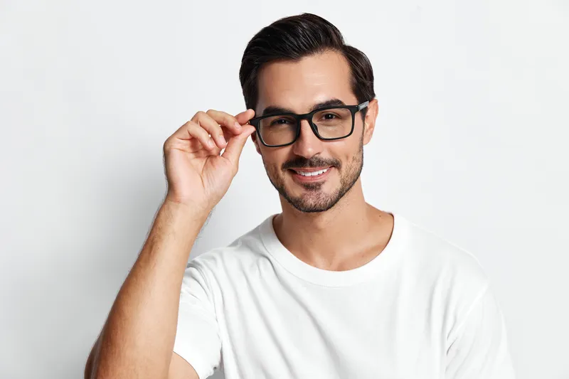 Smiling happy cheerful young man wearing glasses, looking at the camera while adjusting his frames, isolated on plain solid white background. Lifestyle concept.