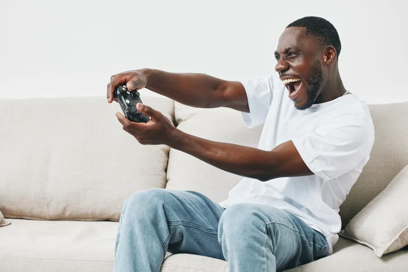 Happy young African American man enjoying gaming on the sofa with a video game controller in hand, expressing excitement and joy in a modern living room