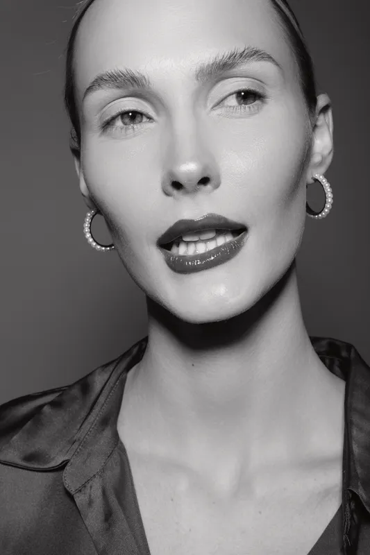 Close-up portrait of a fair skinned white woman with slicked back hair and hoop earrings. Studio lighting emphasizes skin texture and facial expression.
