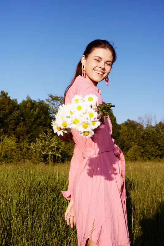 Young woman in a pink dress holding daisies in a sunny field, smiling with joy in the fresh air, showcasing a vibrant and cheerful summer atmosphere