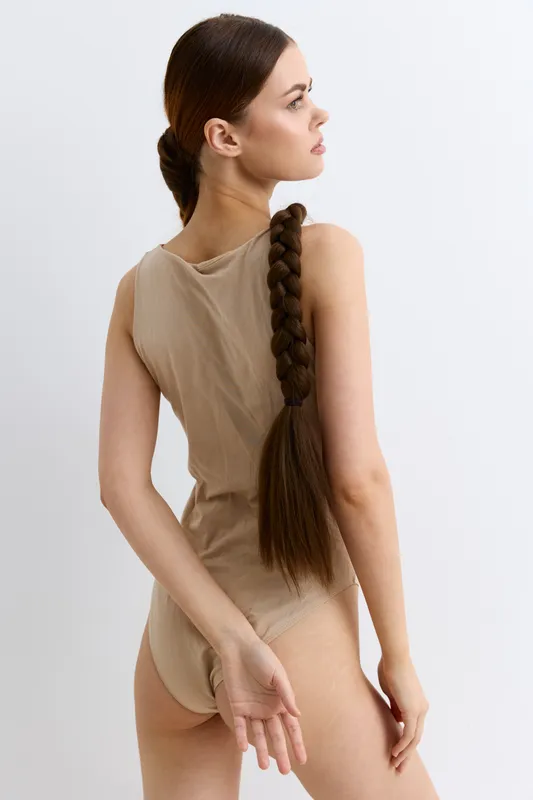 Young woman with long braided hair wearing beige bodysuit poses sideways on white background, showcasing natural beauty and minimalistic style in studio setting.