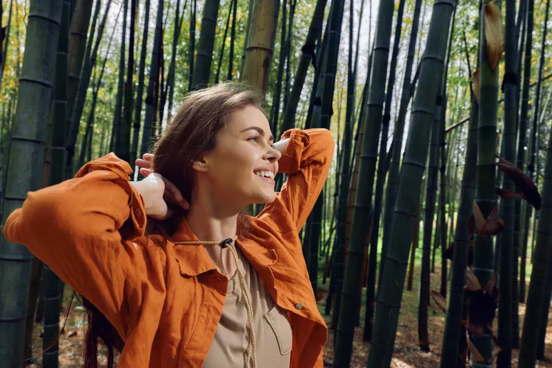 Woman in orange jacket smiling in bamboo forest, portrait shot capturing nature, travel mood and relaxed happiness. Outdoor scene with sunlight, calm expression and joy.