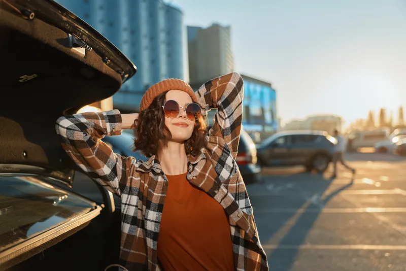 Woman, car, parking, sunset, sunglasses, smile, beanie, urban relaxed young woman leaning on open trunk at golden hour glow, candid lifestyle portrait with authenticity and emotional storytelling.