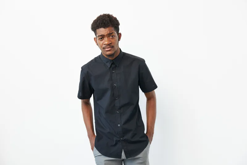 A stylish young African American man in a black shirt stands confidently against a white background, showcasing modern fashion and an approachable demeanor.