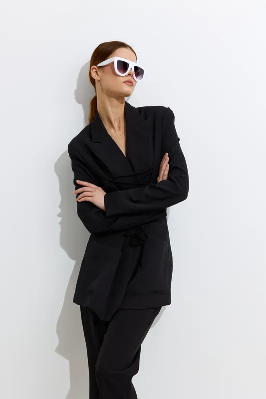 Stylish woman in black suit wearing white sunglasses stands confidently with arms crossed against white background, showcasing modern fashion and minimalistic elegance.