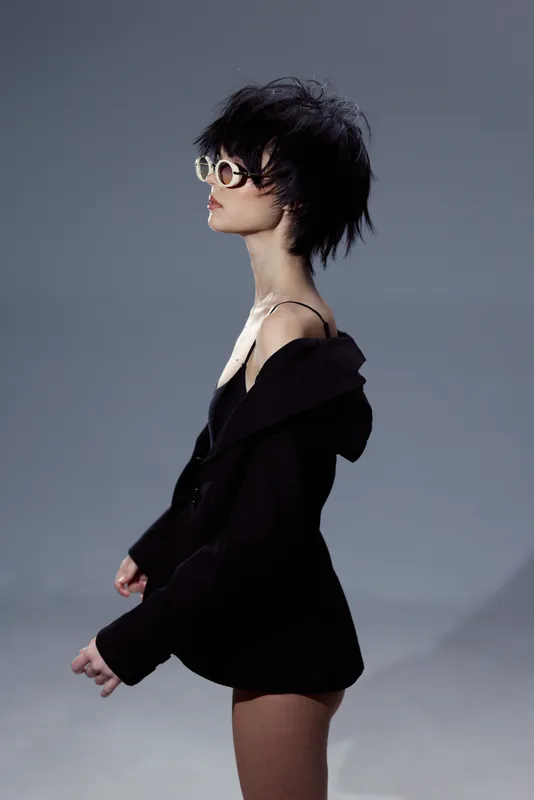 Stylish woman wearing glasses and black jacket posing against gray background with moody expression.