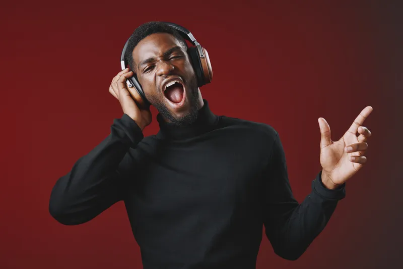 Young Black man enjoying music with headphones, expressing joy and excitement, wearing a black turtleneck against a red background