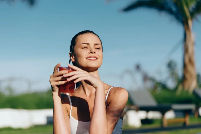 Smiling woman holding a glass outdoors under bright sun, enjoying a refreshing drink. Casual lifestyle scene with warm light, relaxed pose, and a sense of wellness on a sunny day.