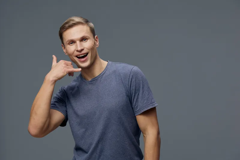smiling young caucasian man casual gray t-shirt show phone gesture communicate video chat remote digital connection concept