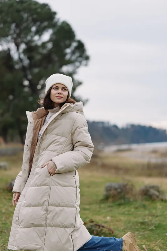 Woman in a beige puffer coat and white beanie standing outdoors in a countryside landscape, winter fashion and cozy style, relaxed pose and natural lighting in chilly weather
