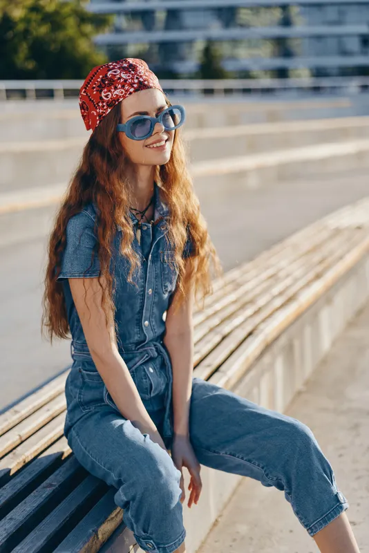lifestyle woman in modern boho-western denim street style wearing sunglasses and red bandana, sitting on bench in bright film color sunlight outdoors