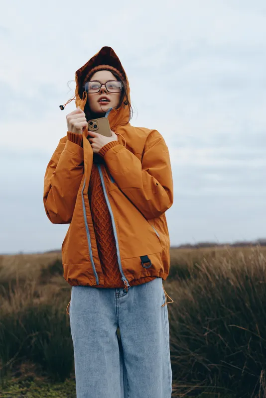 Woman outdoor lifestyle in autumn field wearing hood, jacket, scarf and glasses, smiling as wind lifts her hood, grassy countryside under soft daylight, casual travel