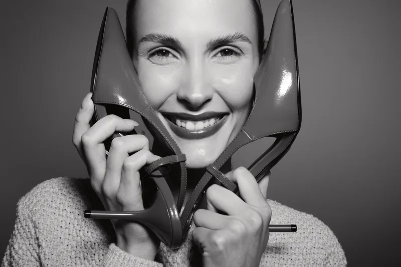 Smiling young white woman holding high heel shoes near face in black and white studio portrait. Concept of fashion, footwear, and happiness.