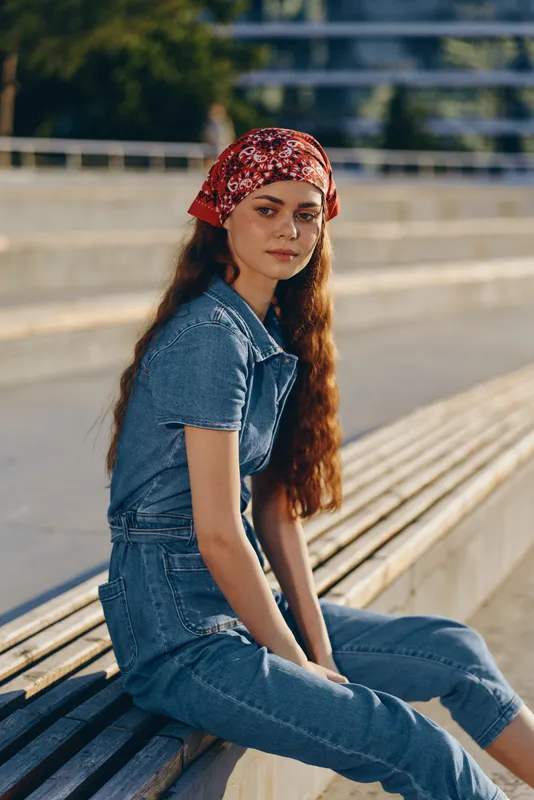 Lifestyle woman wearing modern boho-western denim street style and red bandana, sitting outdoors in warm natural light with film color effect and relaxed mood.