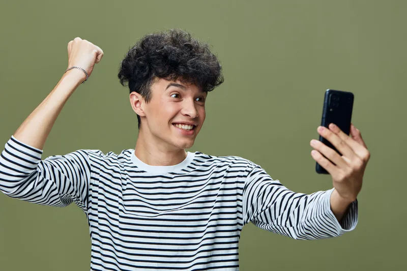 excited young man holding smartphone celebrating victory with fist pump wearing striped shirt and curly hair against green background
