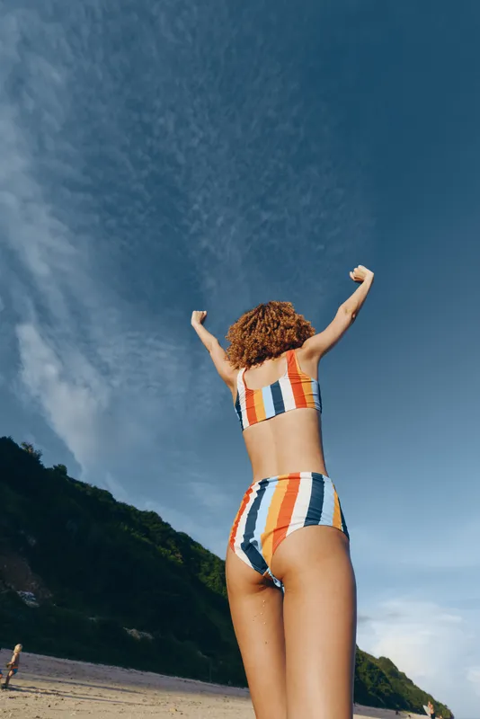 A woman in a striped bikini stands on the beach with arms raised, celebrating sunshine and freedom. Clear blue sky, sandy shore, and distant cliffs create a vibrant summer scene.